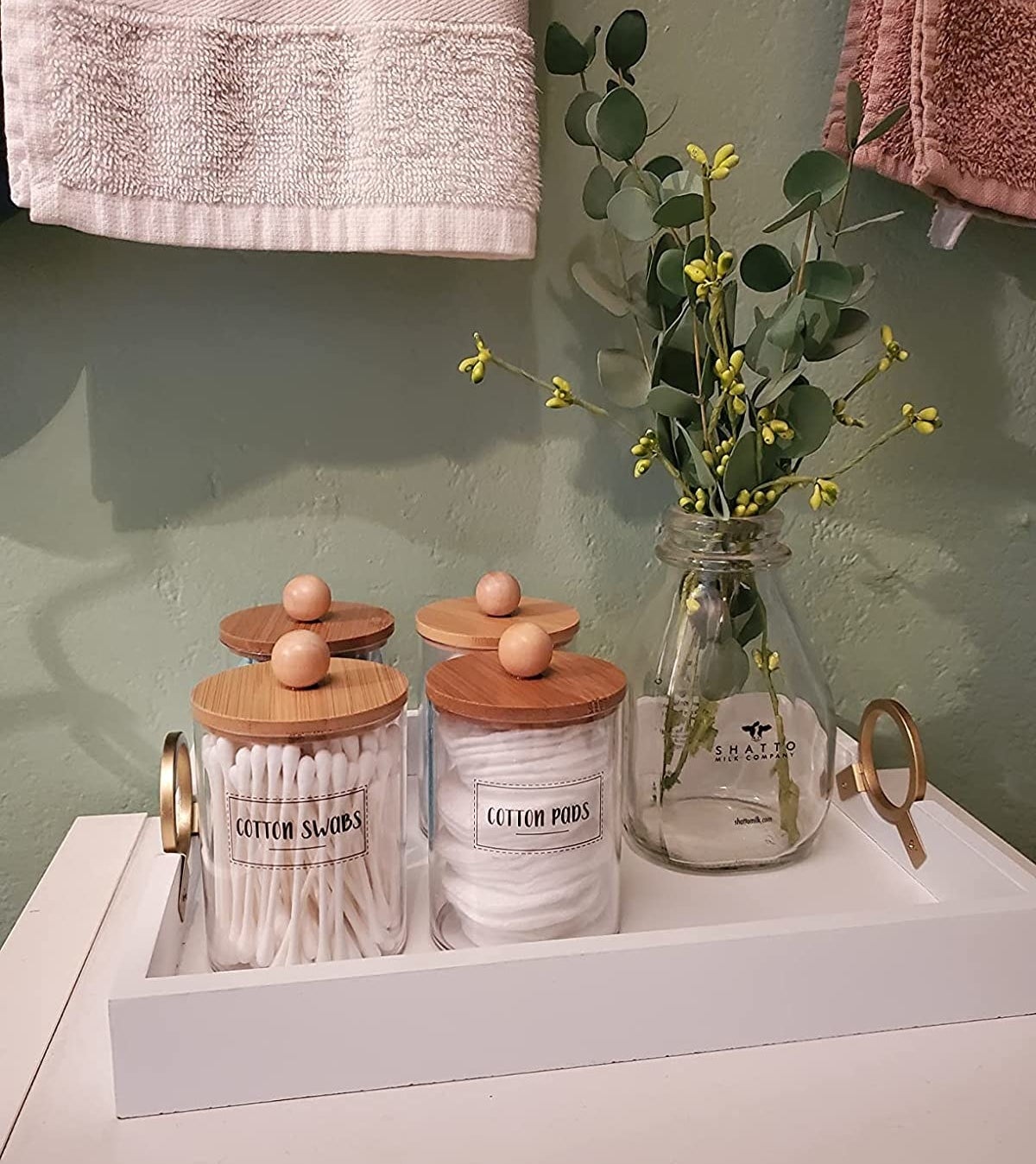 a reviewer photo of the labeled glass jars with wood lids filled with q-tips and cotton pads on a tray with a vase of eucalyptus