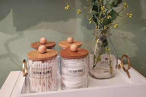a reviewer photo of the labeled glass jars with wood lids filled with q-tips and cotton pads on a tray with a vase of eucalyptus