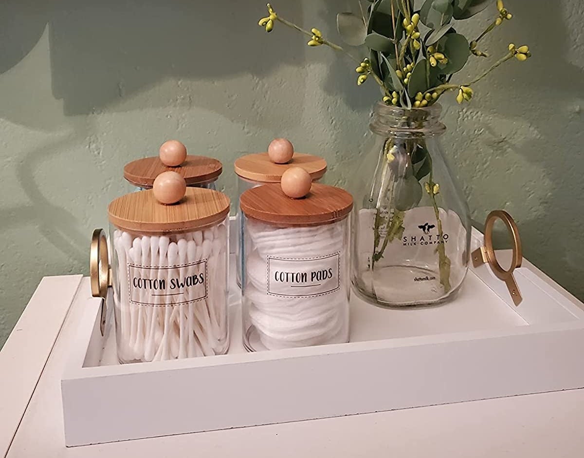 a reviewer photo of the labeled glass jars with wood lids filled with q-tips and cotton pads on a tray with a vase of eucalyptus