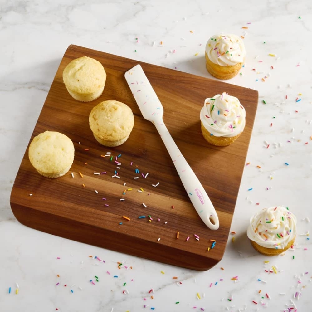 the spatula on a cutting board with cupcakes half of them which are iced and sprinkles everywhere