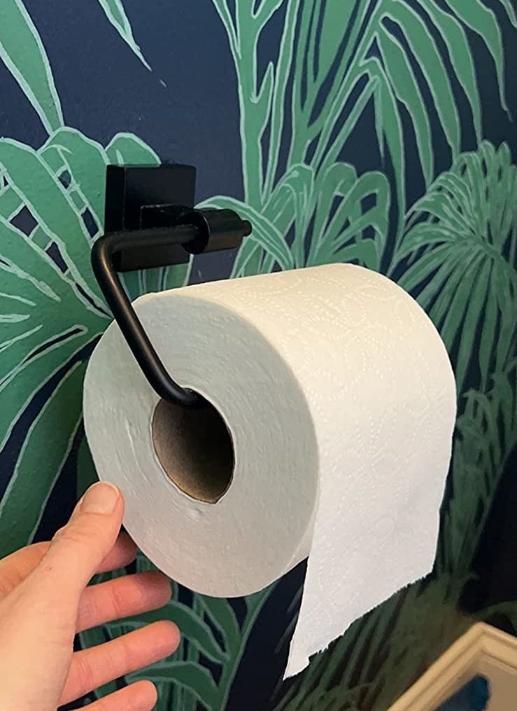 a reviewer photo of a hand next to the matte black toilet paper holder on green and blue wallpaper