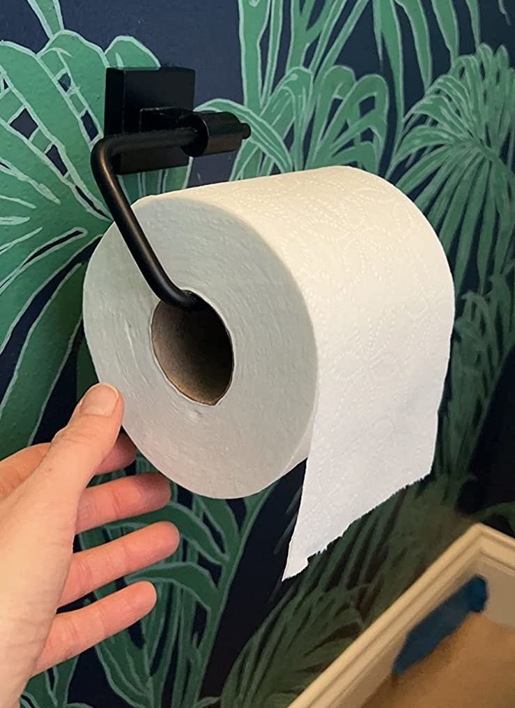 a reviewer photo of a hand next to the matte black toilet paper holder on green and blue wallpaper