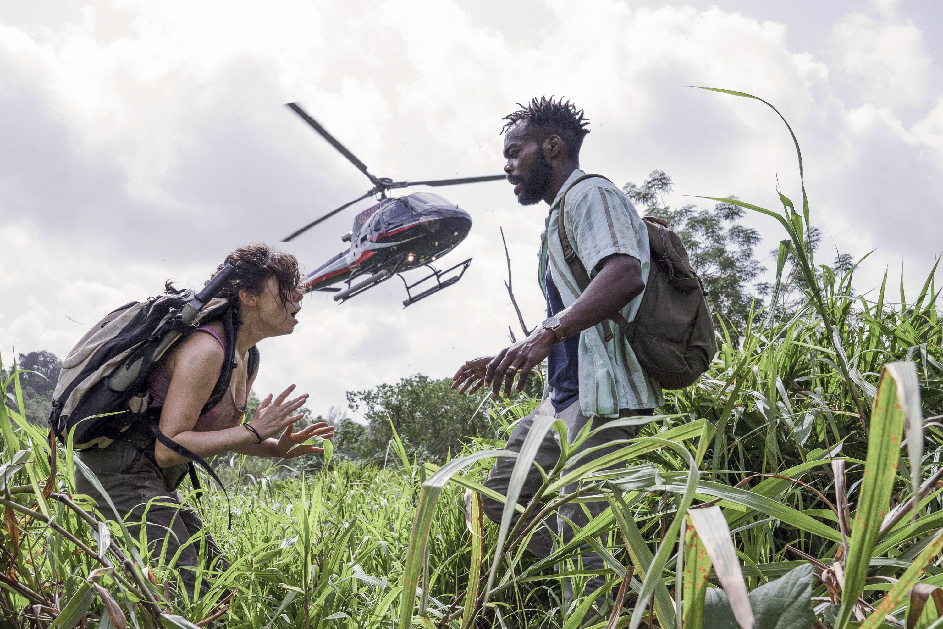 William Jackson Harper and Cristin Milioti stand outside in tall grass. There is a helicopter in the background in The Resort.