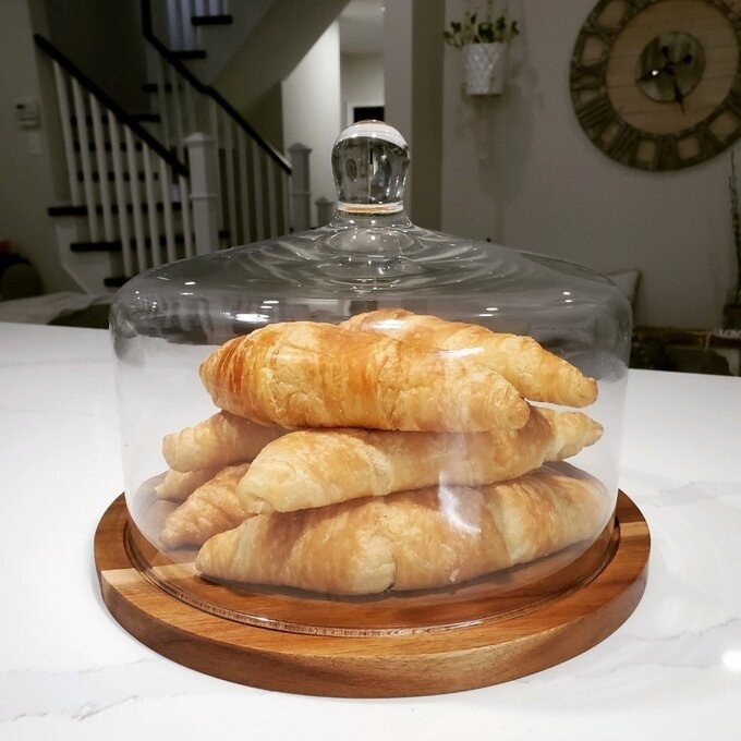 A glass dome on a wooden platter covers croissants on a kitchen counter