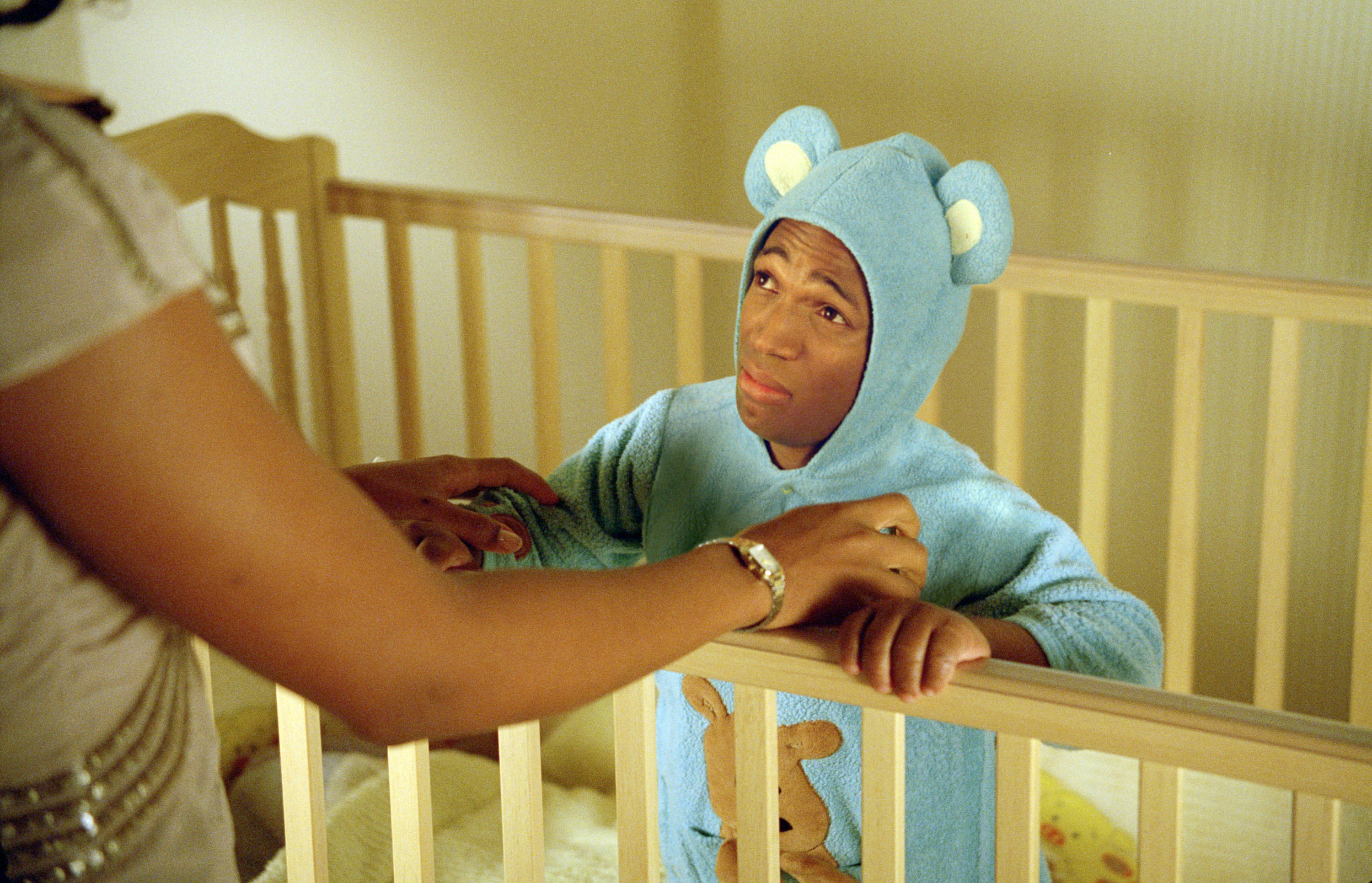 A little person pretending to be an infant winces when forced to wear a teddy bear onesie