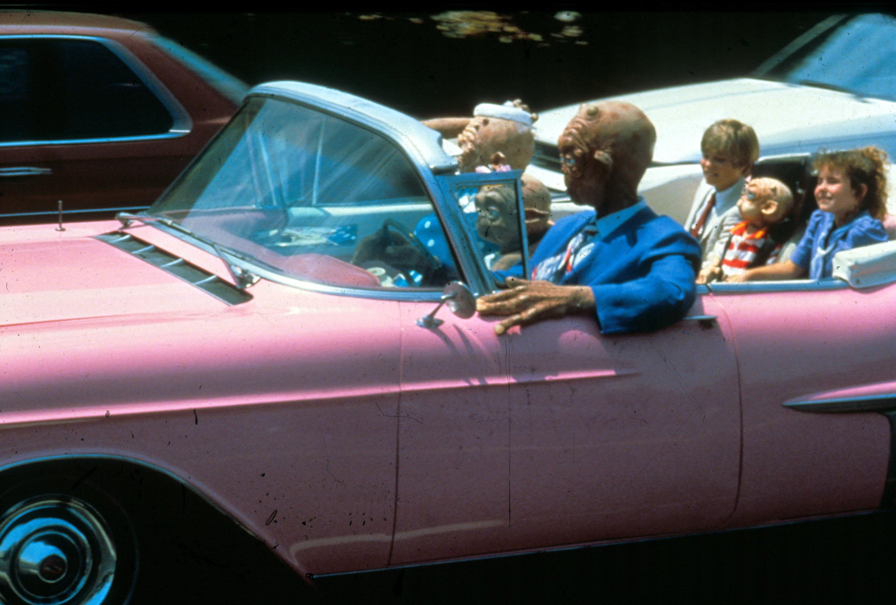 Two kids sit in a pink convertible with a family of formally-dressed aliens