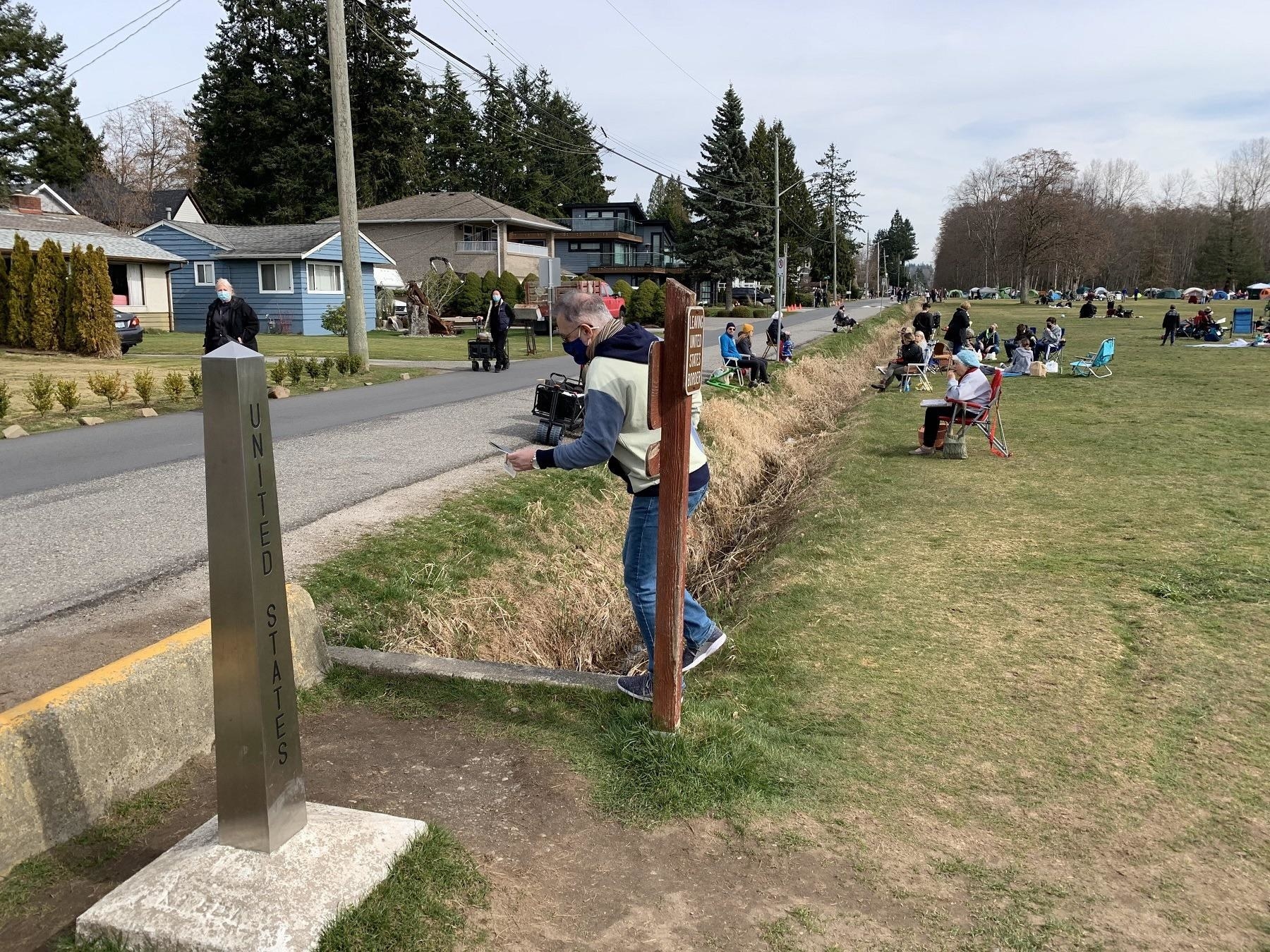 Two poles in the ground separating the US from Canada, with people sitting on the grass on the Canadian side