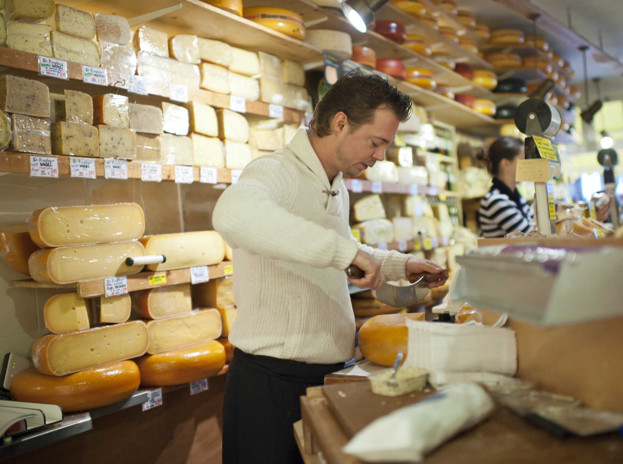 a man cutting a big block of cheese with countless rolls of cheese stacked behind him on shop shelves
