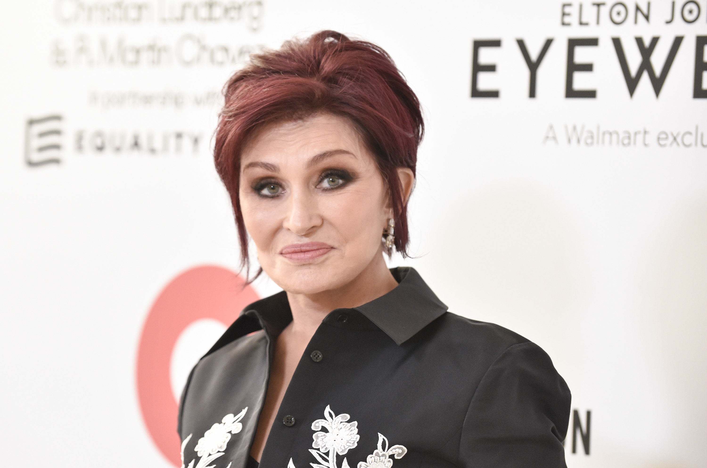 Sharon Osbourne posing at an eyewear event in a black floral button down shirt
