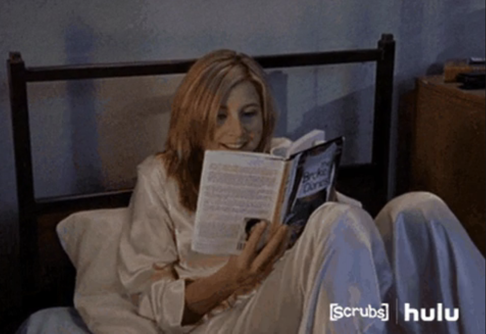 Woman in silky pajamas sitting up in bed and reading a book while smiling