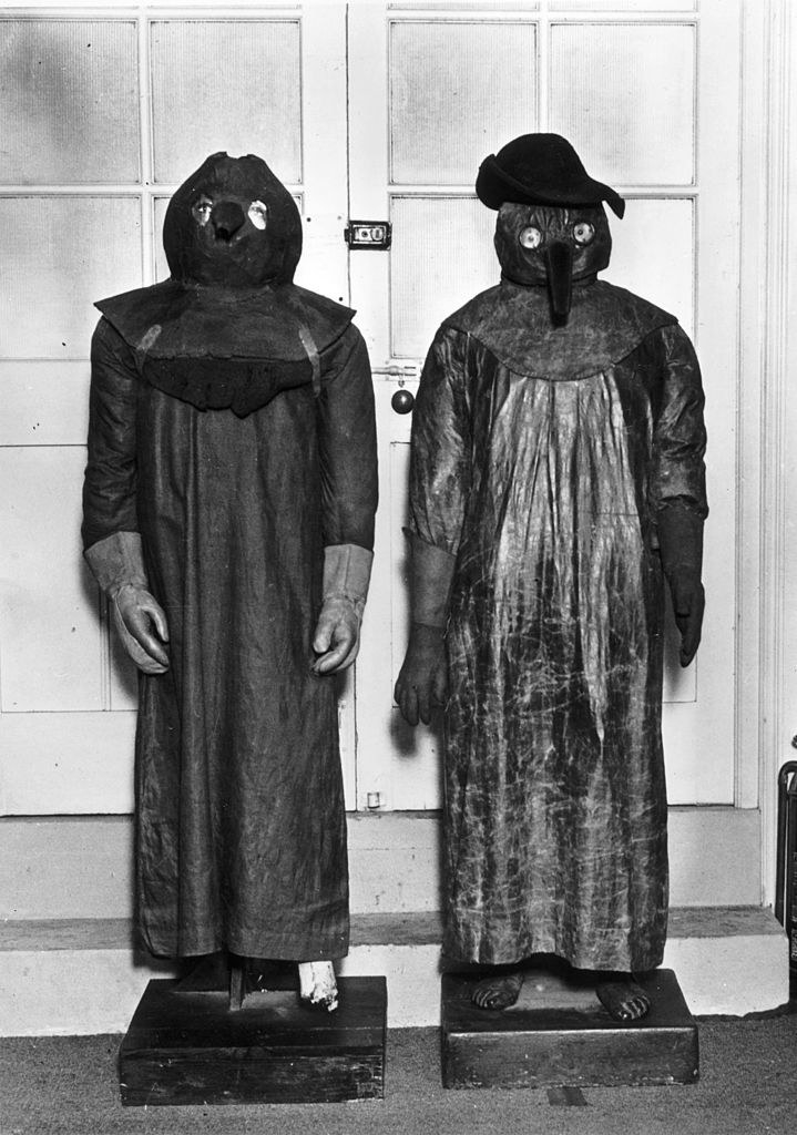 Two people in historical plague-doctor costumes with long robes, long gloves, and beaklike masks that have eye openings and extend to the shoulders