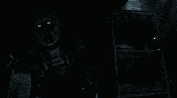 A large-headed monster with glowing eyes walks through a dark basement
