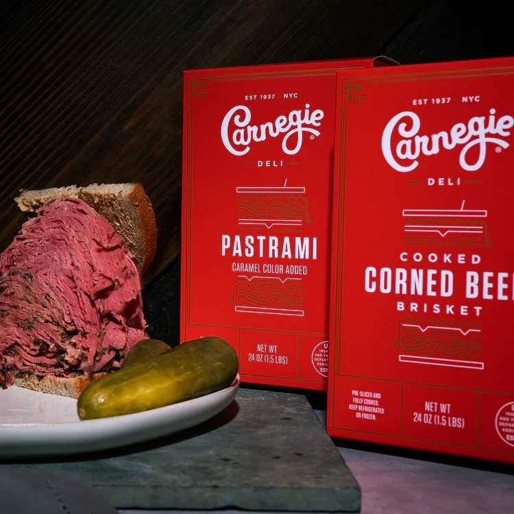Red boxes that say &quot;Carnegie Deli: Cooked Corned Beef Brisket&quot; sitting next to a plate with a corned beef sandwich with a pickle.