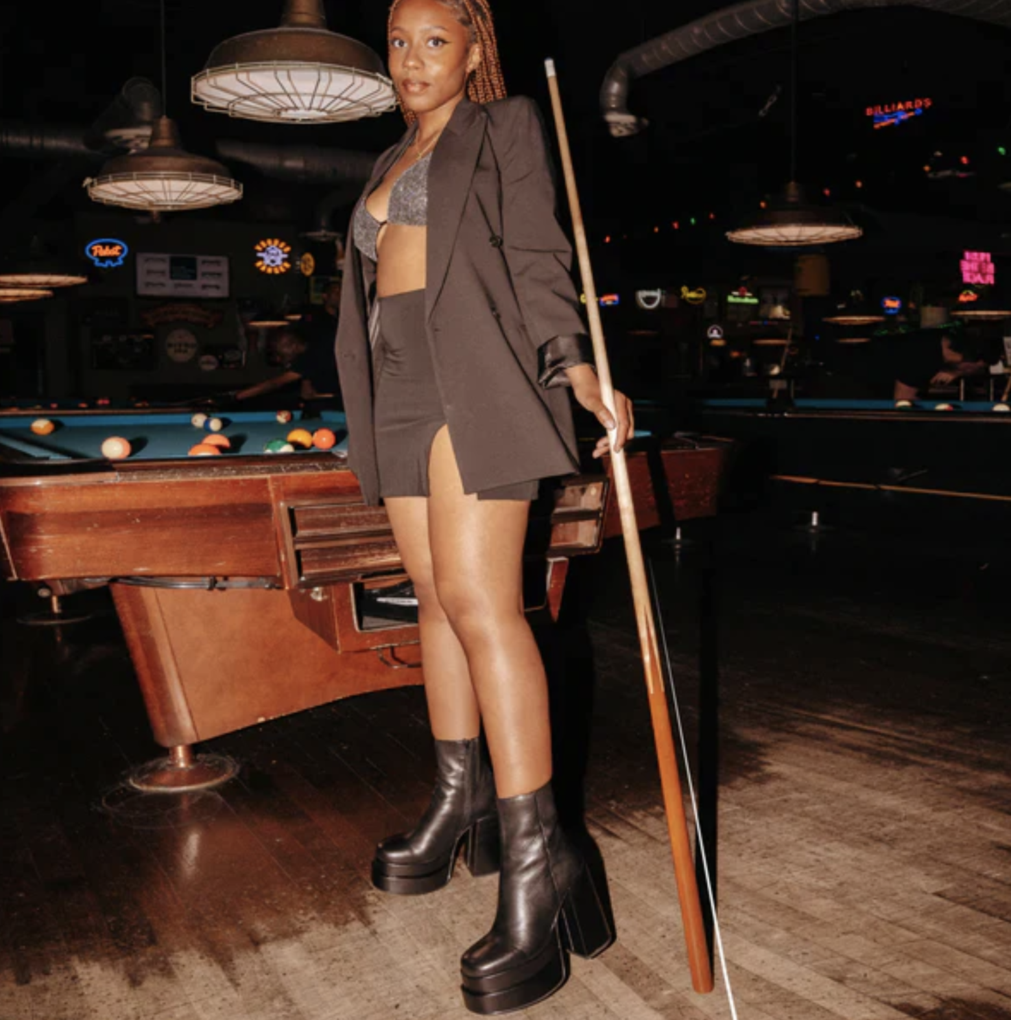 A person wearing the boots with a blazer, top, and skirt while standing at a pool table with a pool cue