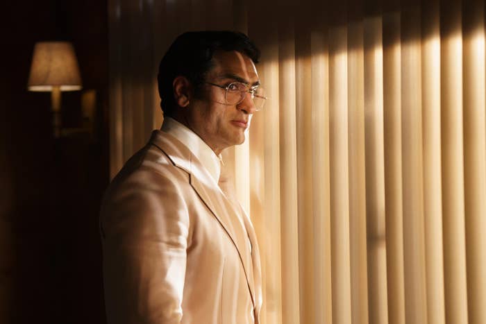 Kumail as Somen standing in front of a sliding glass doors covered with blinds