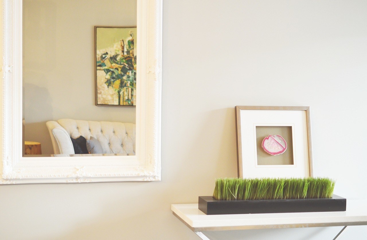 A white bordered mirror against a plain white wall with a plant and frame on the right on a table
