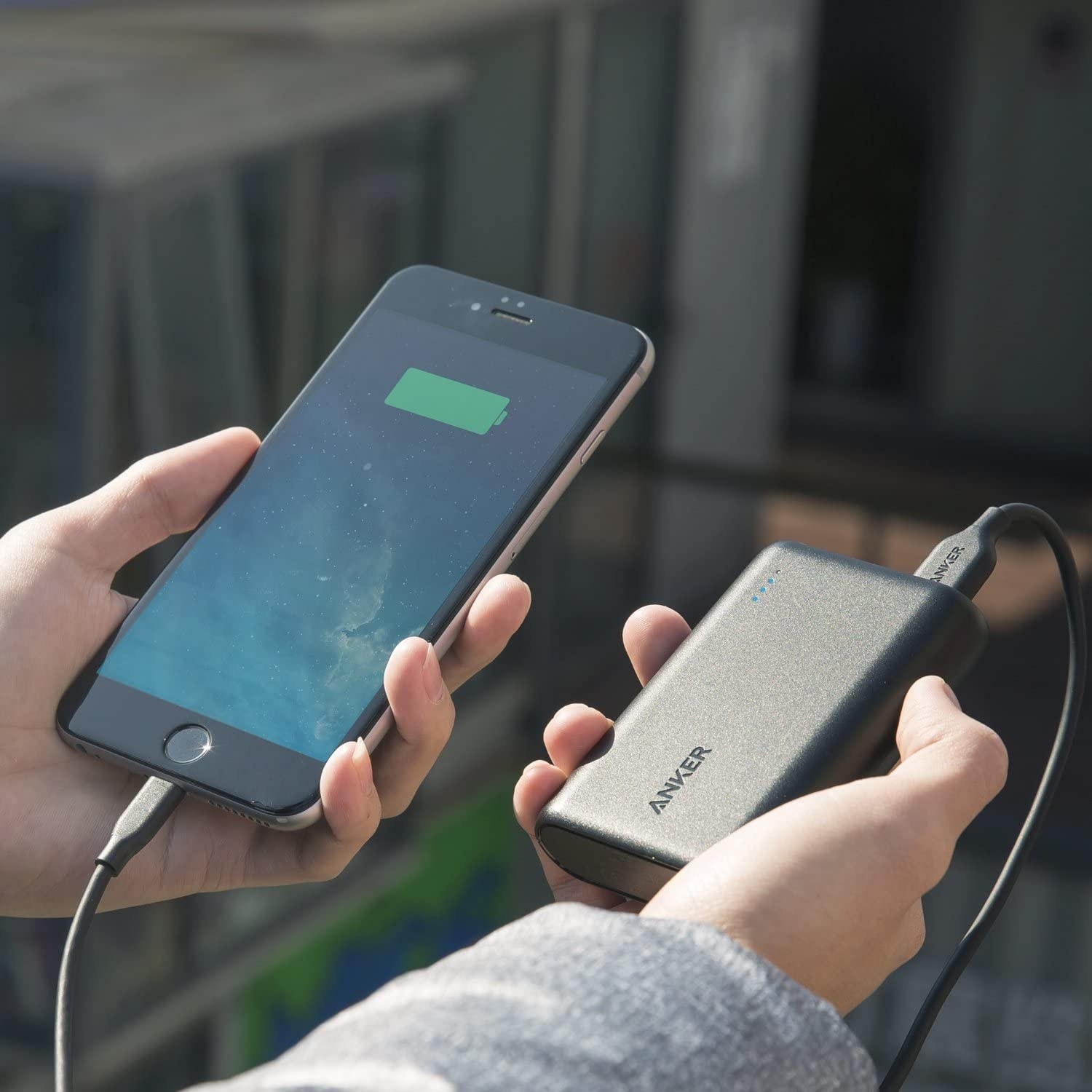 Two hands holding a smartphone being charged by a portable charging device via USB cable
