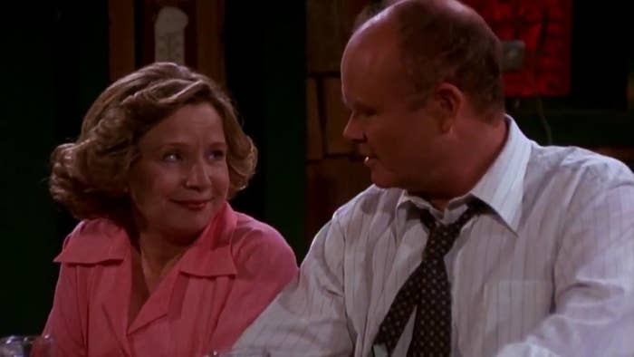 Kitty and Red look at each other, talking about financial hardships. Kitty is wearing a pink shirt, and Red is in a button up with a disheveled tie.