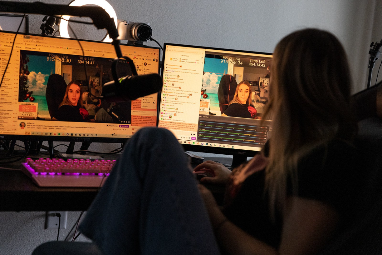 A woman with long hair looks at two computer screens featuring videos of herself