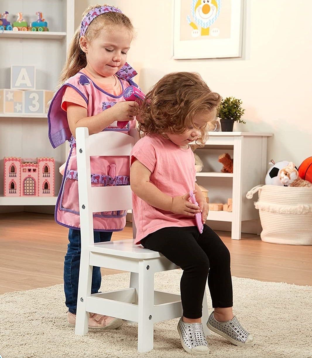 One kid pretending to cut another kid&#x27;s hair as they sit on a stool in a playroom