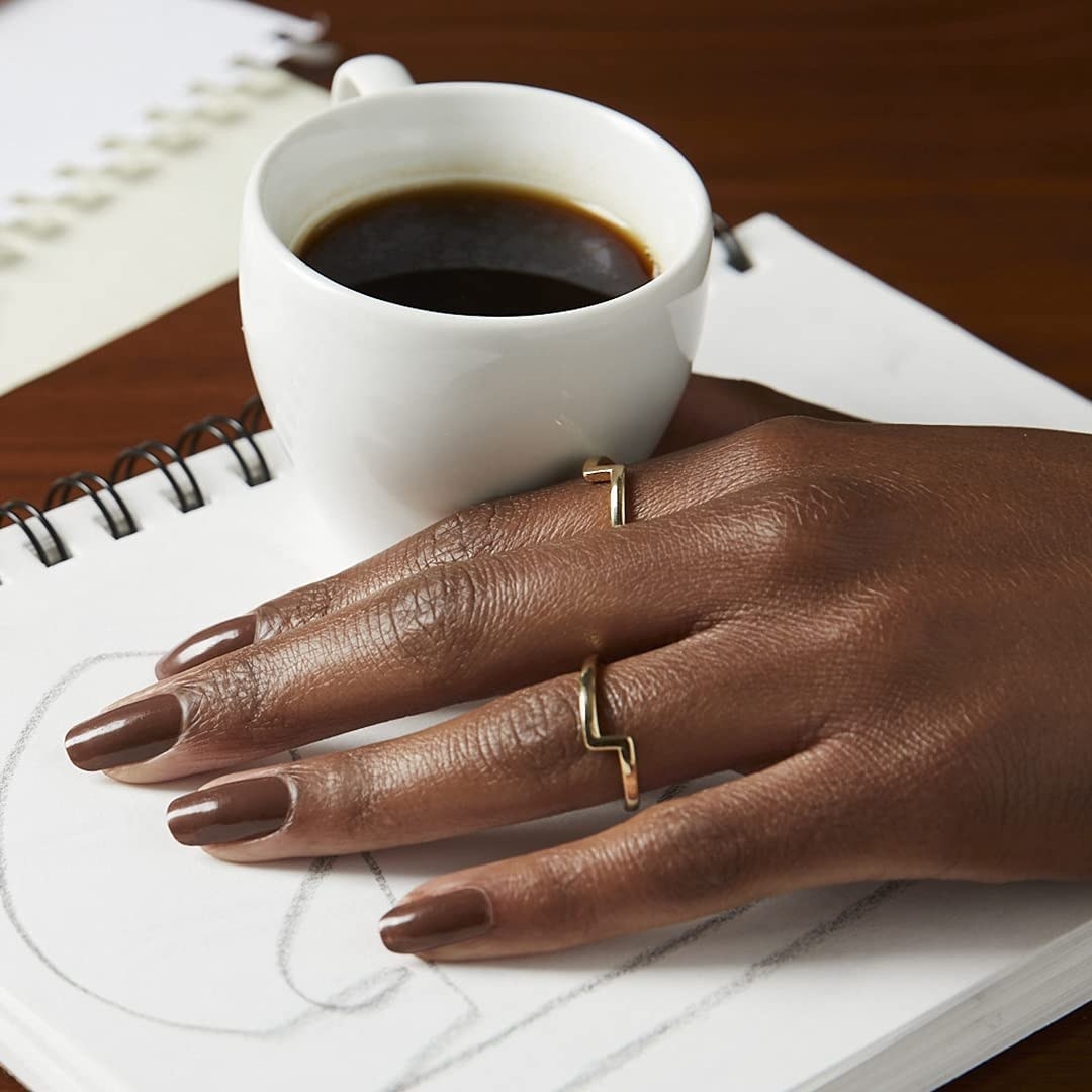 A person with their nails painted and their hand on a sketchbook beside a cup of coffee