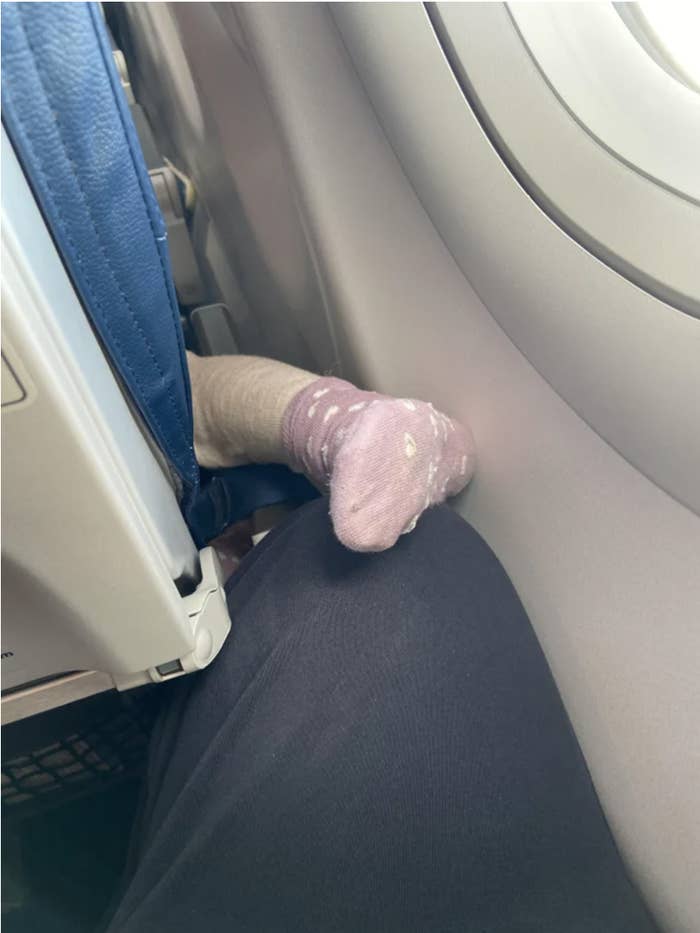 A toddler&#x27;s socked foot and leg protruding from the side of an airplane seat and  almost resting on an adult&#x27;s knee