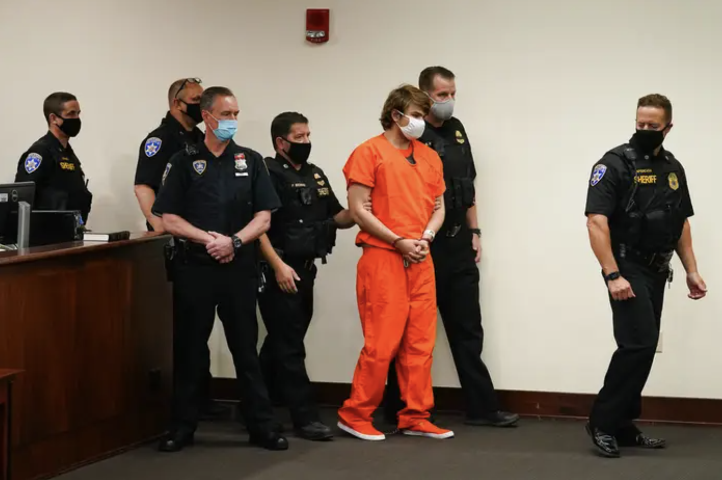 A young man in an orange prison jumpsuit walks through a court room flanked by police officers