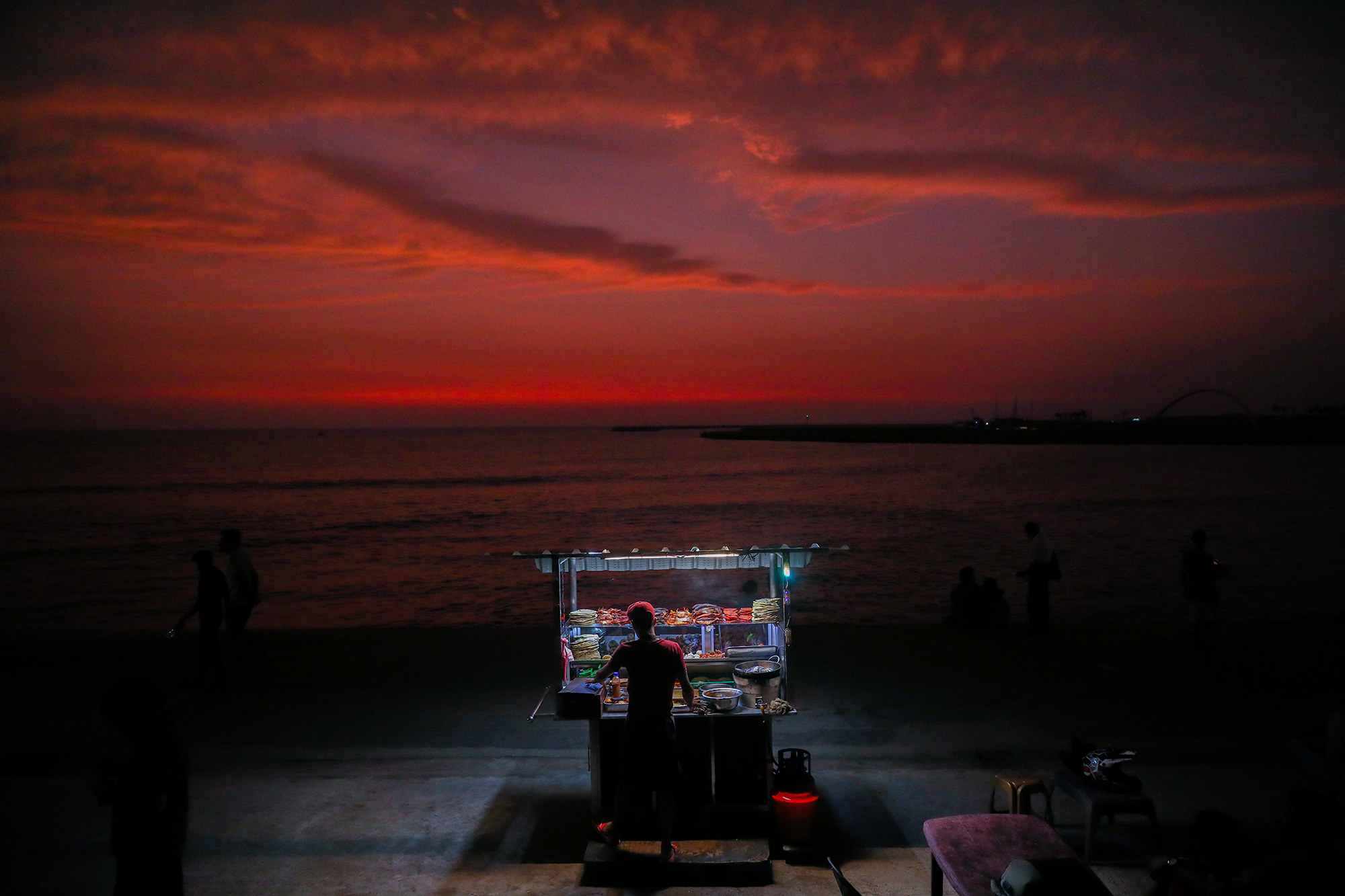 A dark red sunset on the water; in the foreground, the outline of a street vendor is lit up by his food cart