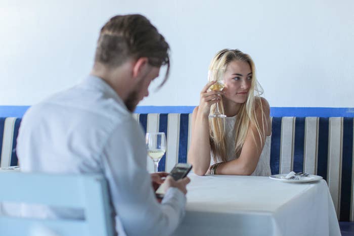 A man and woman at dinner, with him on his phone and her drinking wine