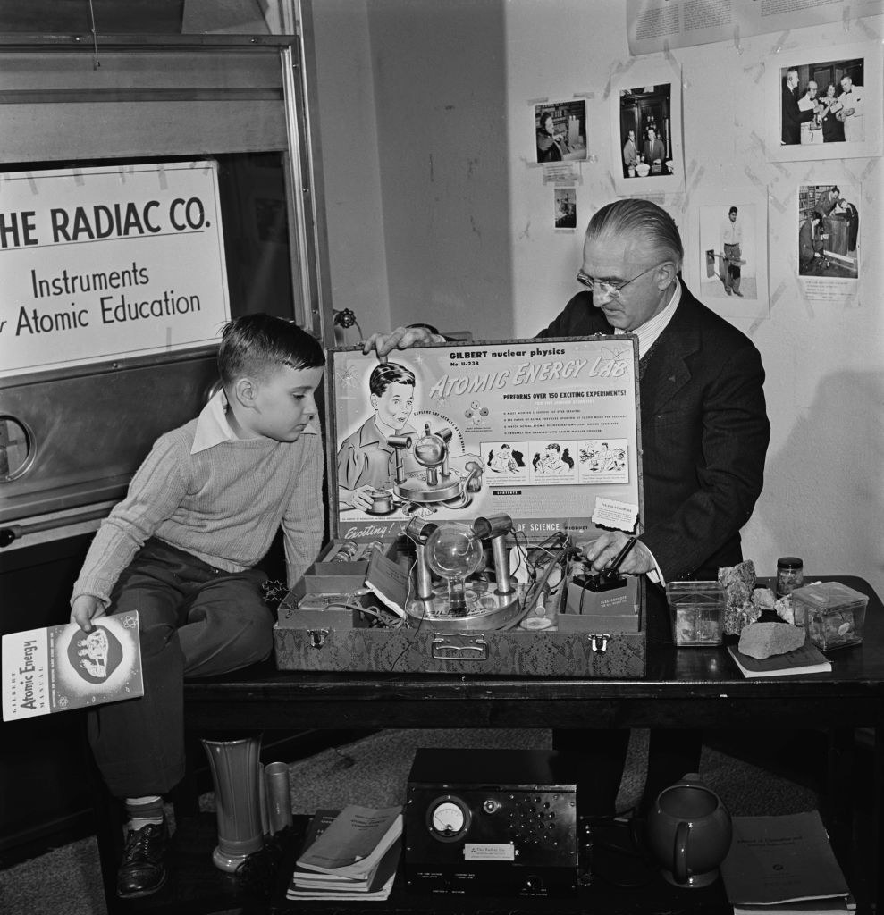 A black-and-white photo of a young boy sitting on a table alongside the &quot;toy,&quot; which appears inside a suitcase held open by an older man and contains a battery-powered Geiger–Müller counter and a &quot;cloud chamber&quot; with radioactive samples