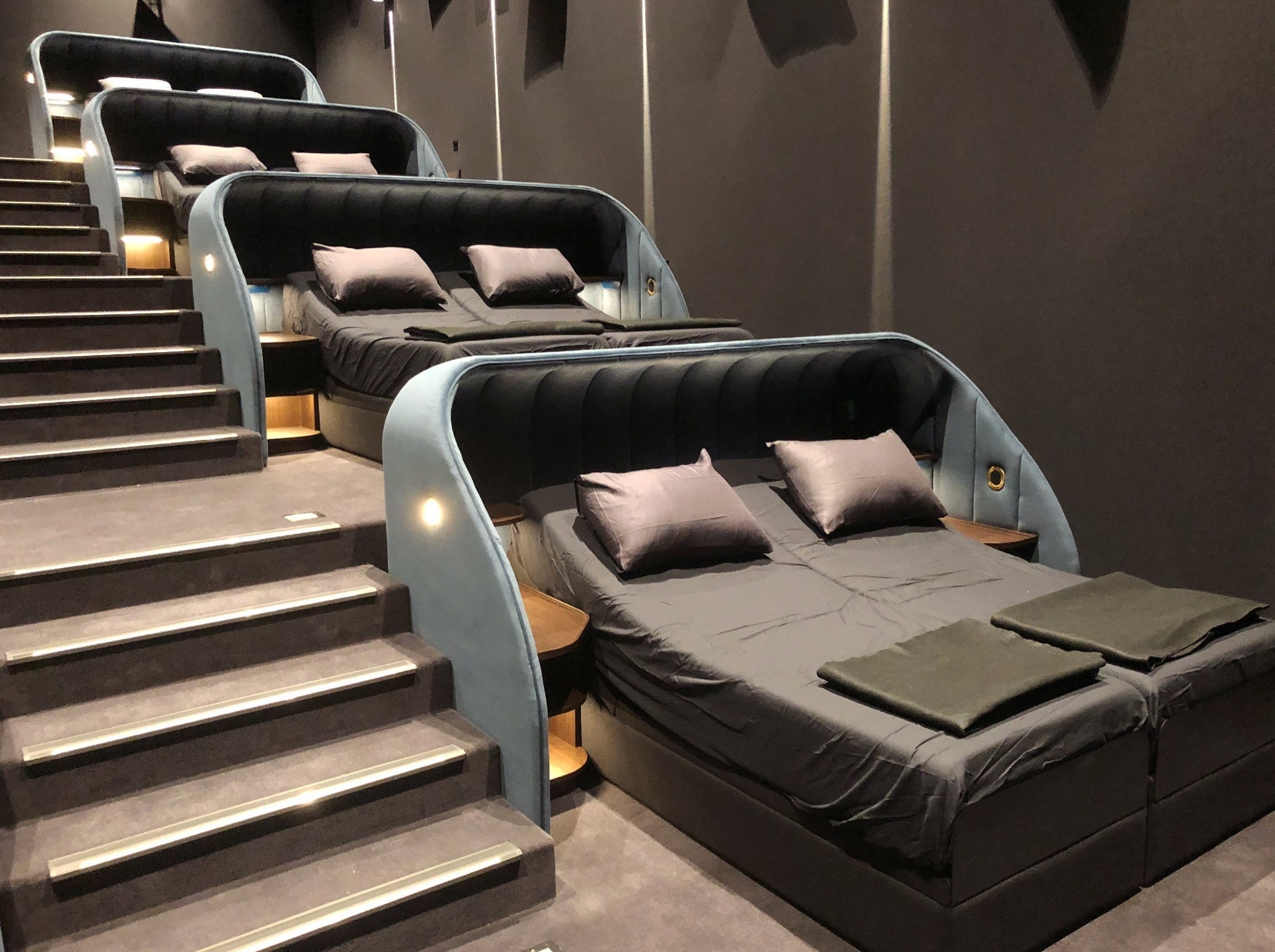 A movie theater with a row of side-by-side twin beds with the heads raised — and including pillows, folded sheets, and small end tables — inside their separate enclosures