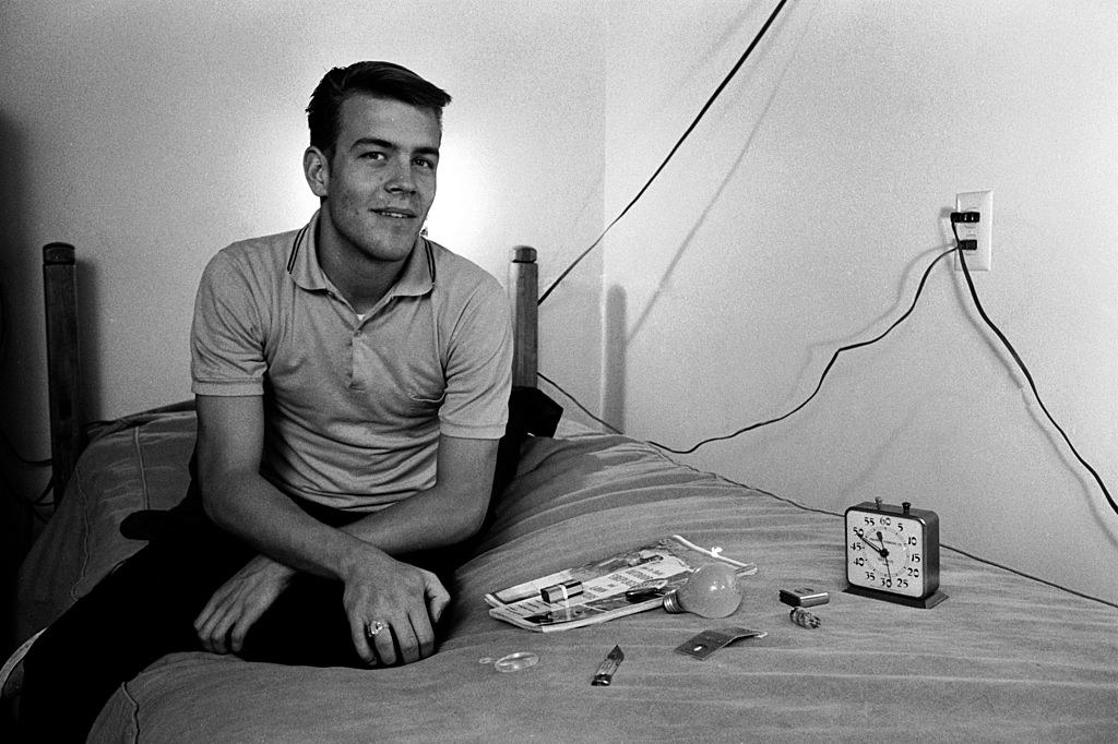 A young man looking remarkably alert sits on a bed with a clock, light bulb, and other items on it