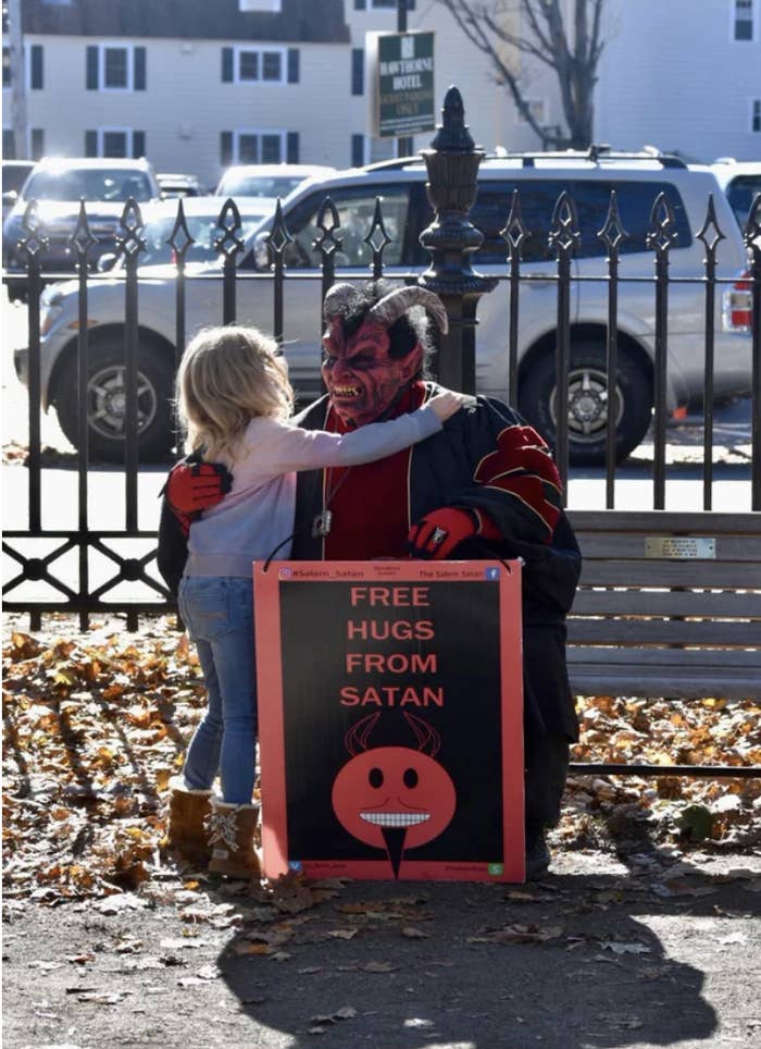 Little blonde girl hugging a man in a Satan costume in front of a sign &quot;Free hugs from Satan&quot;