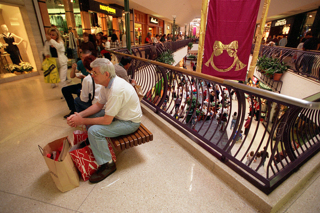 People in a mall, including a man sitting with bags and looking morose