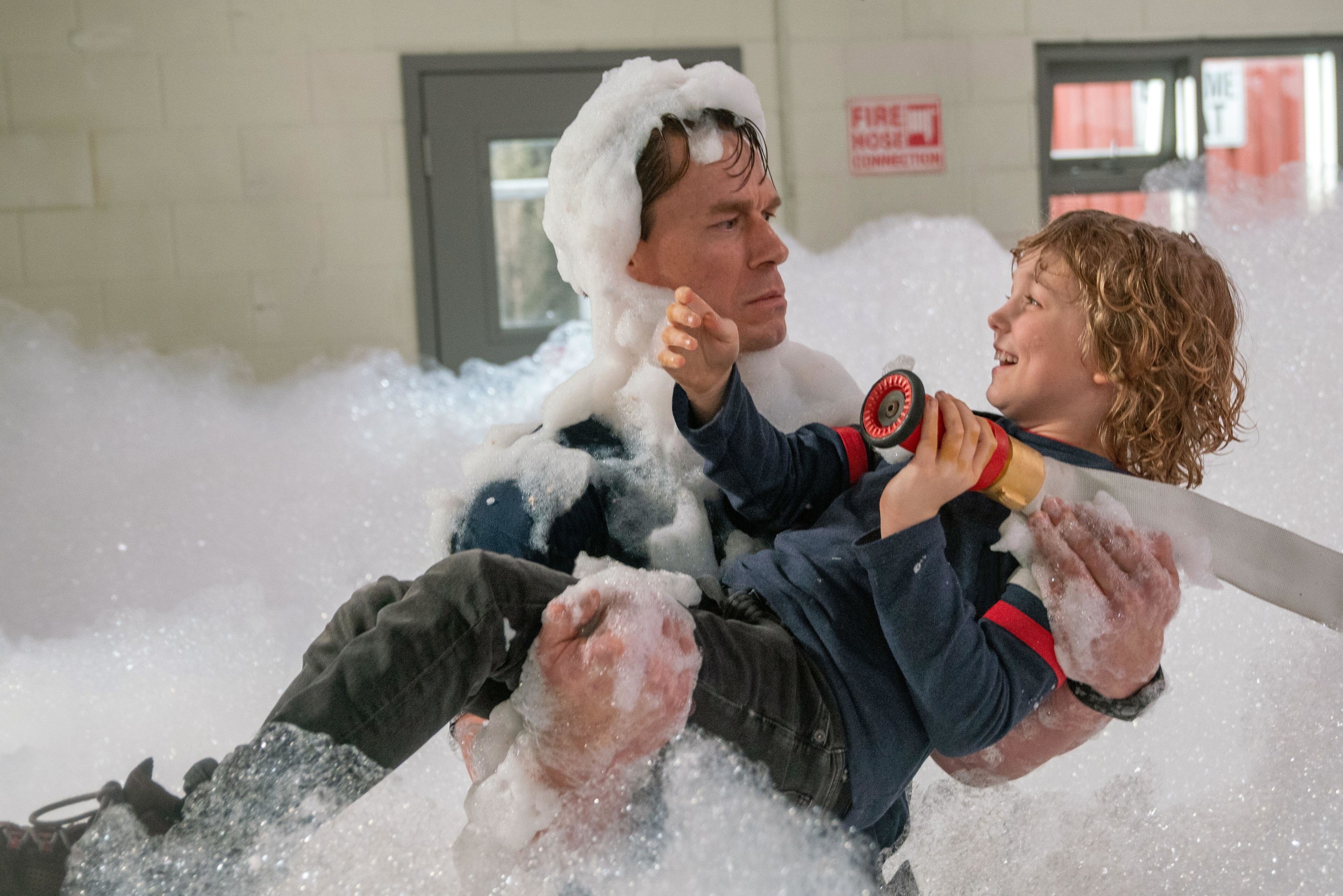 A disappointed man holds a smiling child in a room filled with foamy bubbles