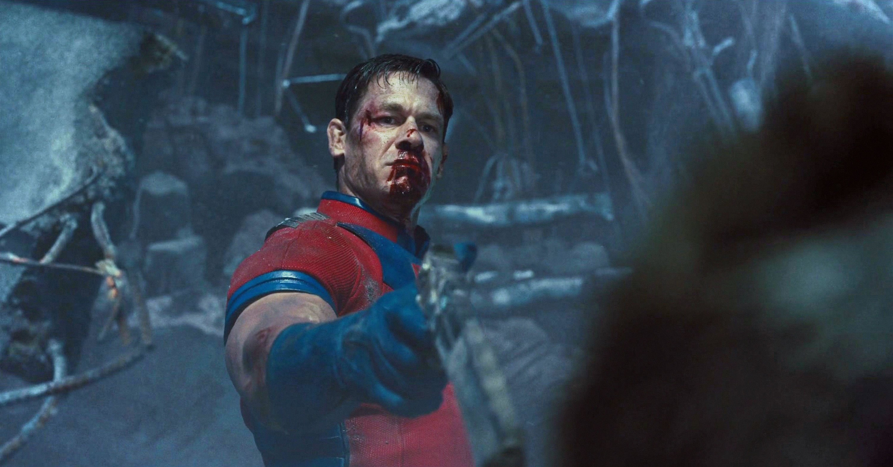A bloodied man in a red-and-blue costume holds a pistol in the wreckage of a building
