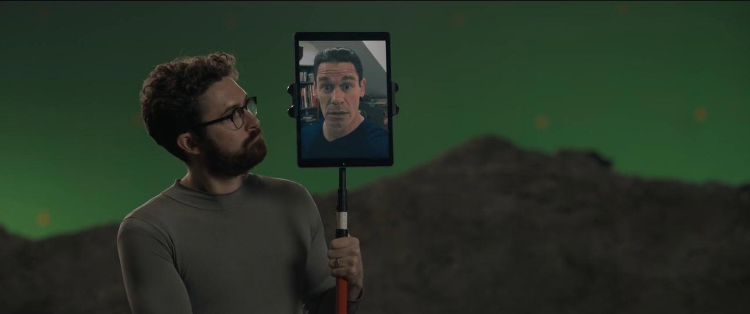 A bearded man with glasses points an tablet with a man&#x27;s face on it against a green screen background