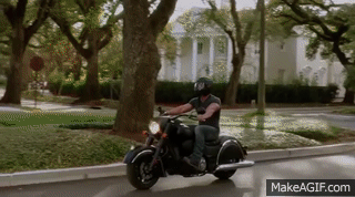 A muscular man in a motorcycle pulls into a driveway and removes his helmet safely.