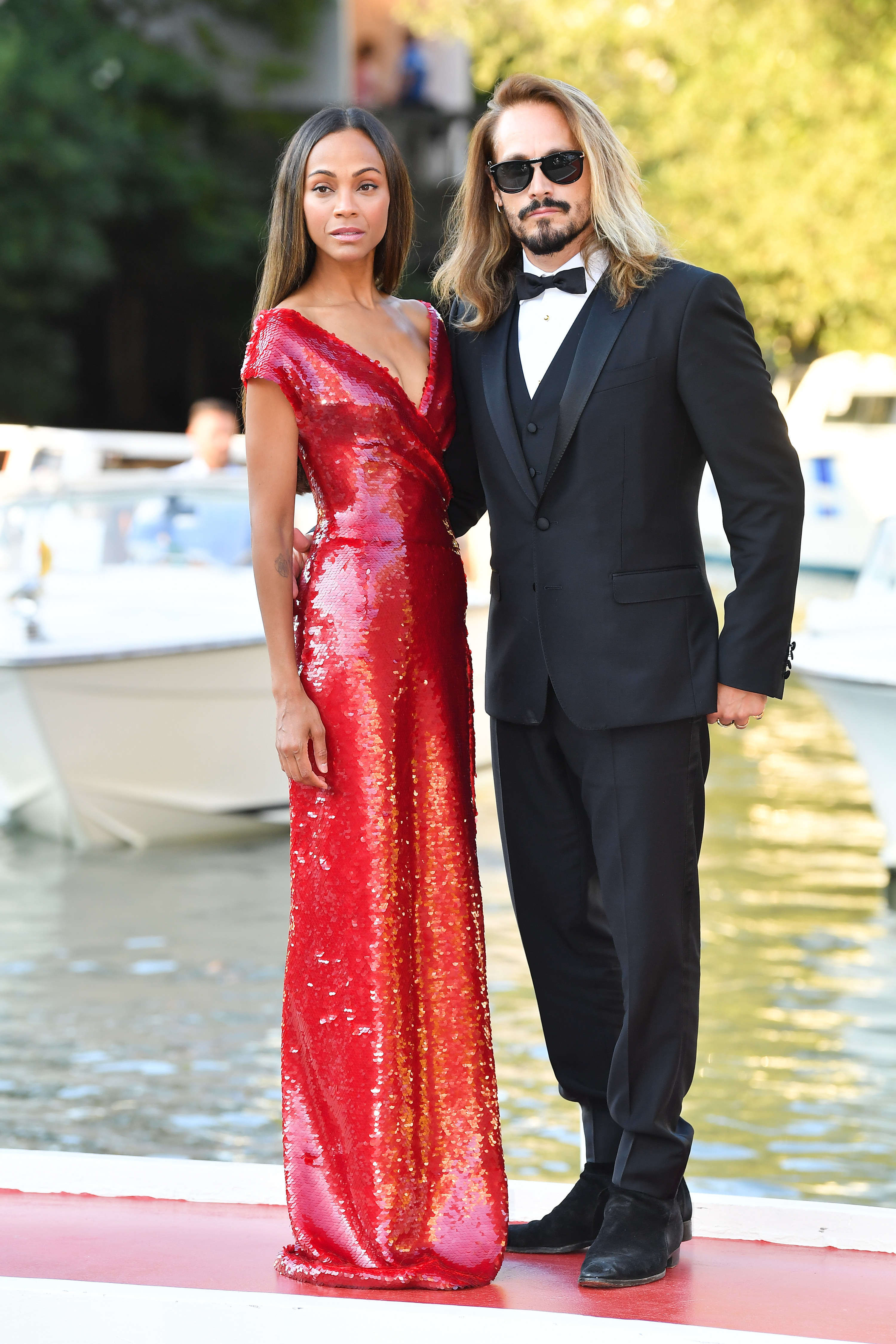 Zoe, wearing a long sequined gown, and Marco, wearing a tux, pose for photographers at an event. with boats behind them