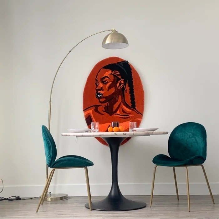 the arched gold floor lamp between two green chairs, a table, and an orange tapestry of a woman with a braid