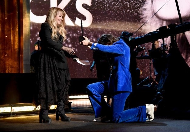 Harry Styles kneels in front of Stevie Nicks and hands her an award