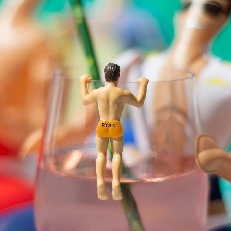 the small man-shaped drink marker in orange shorts with &quot;ryan&quot; on the butt hanging from the edge of a wine glass