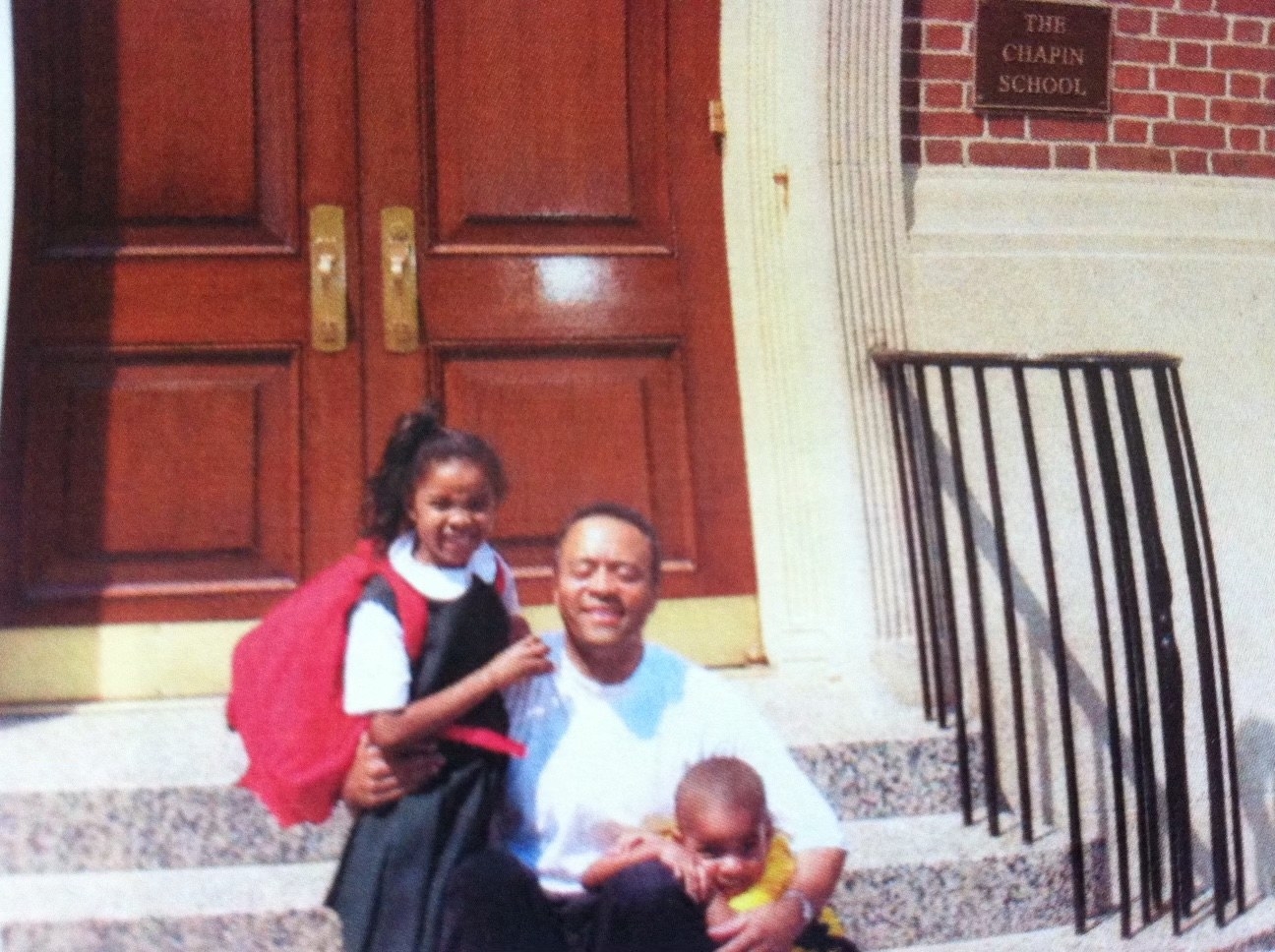 Gabrielle, young and in a school uniform with a big red backpack, in the arms of her uncle that sits on the sets of a school building. Her brother is under his other arm in a yellow shirt.