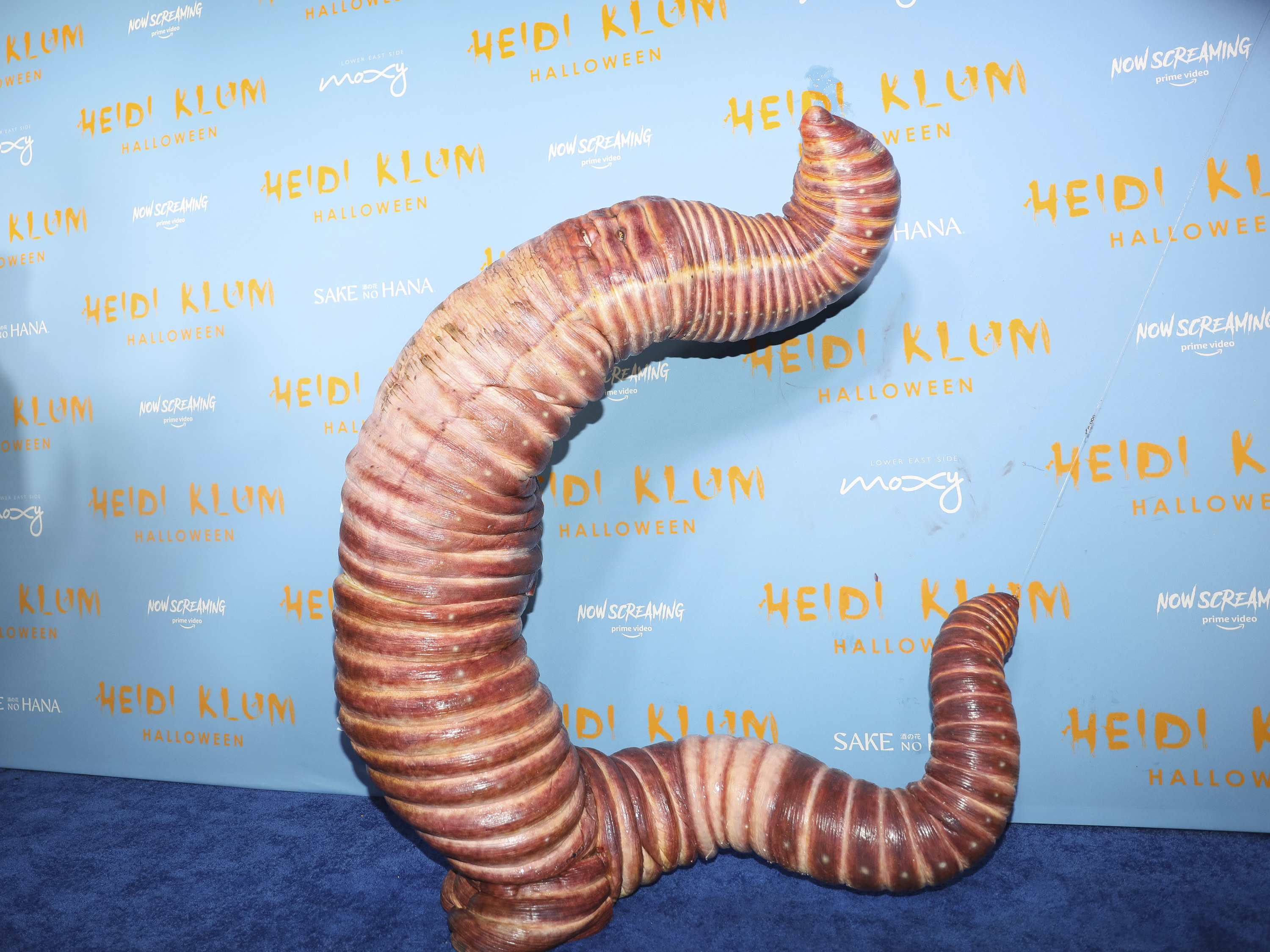 A giant worm costume stands, leaning back on a blue carpet
