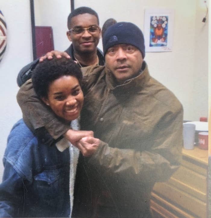 Uncle Blakey, a Black man, with Gabrielle under his arm. She is smiling. Her brother is behind with his hand on her head