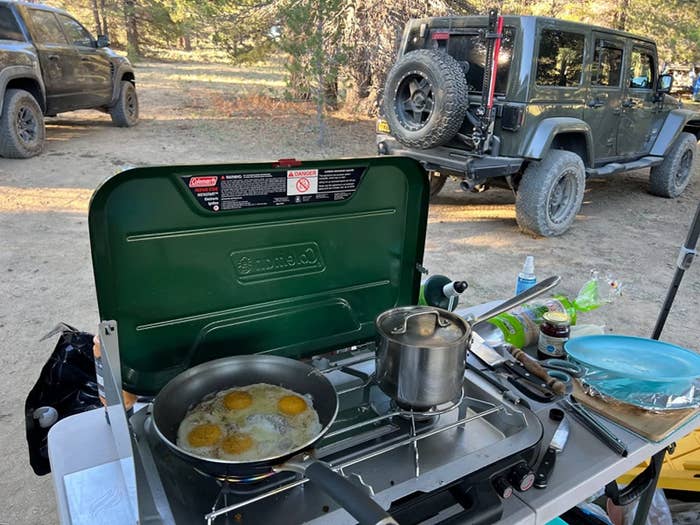 reviewer&#x27;s camp stove at a campsite with a pot on one burner and a pan frying eggs on another
