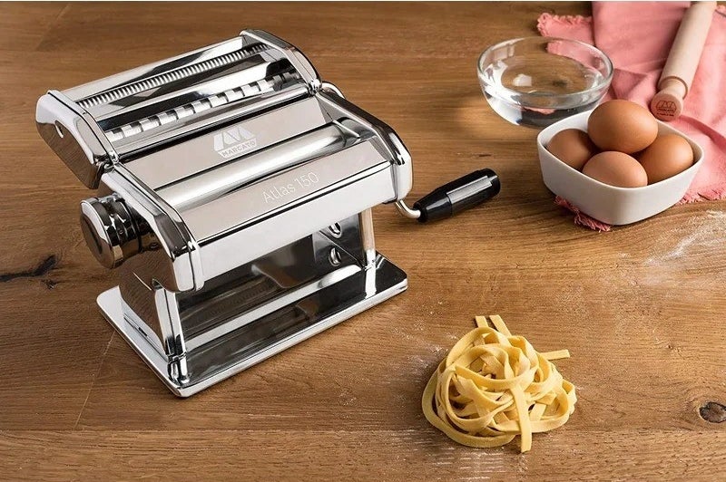 the manual pasta maker next to a bowl of eggs and a pile of noodles