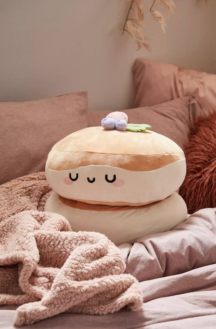 The plushie has a smiling face with closed eyes and is in a light pink bed with various throws and pillows