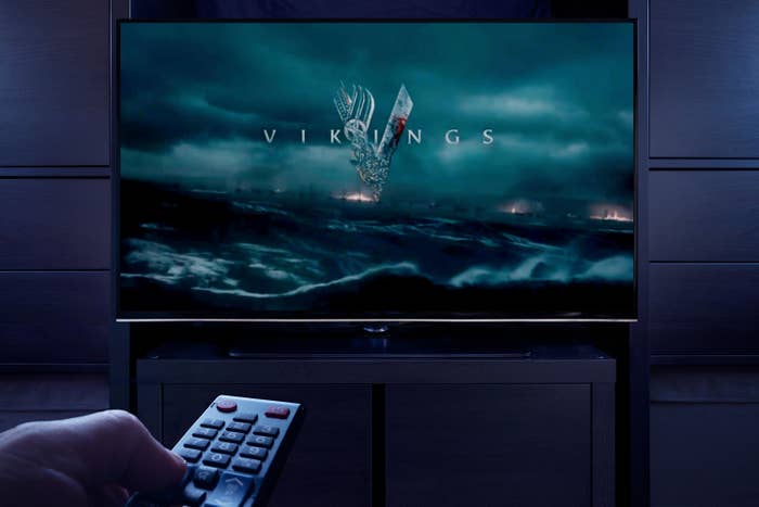 A man points a TV remote at the television which displays the Vikings main title screen