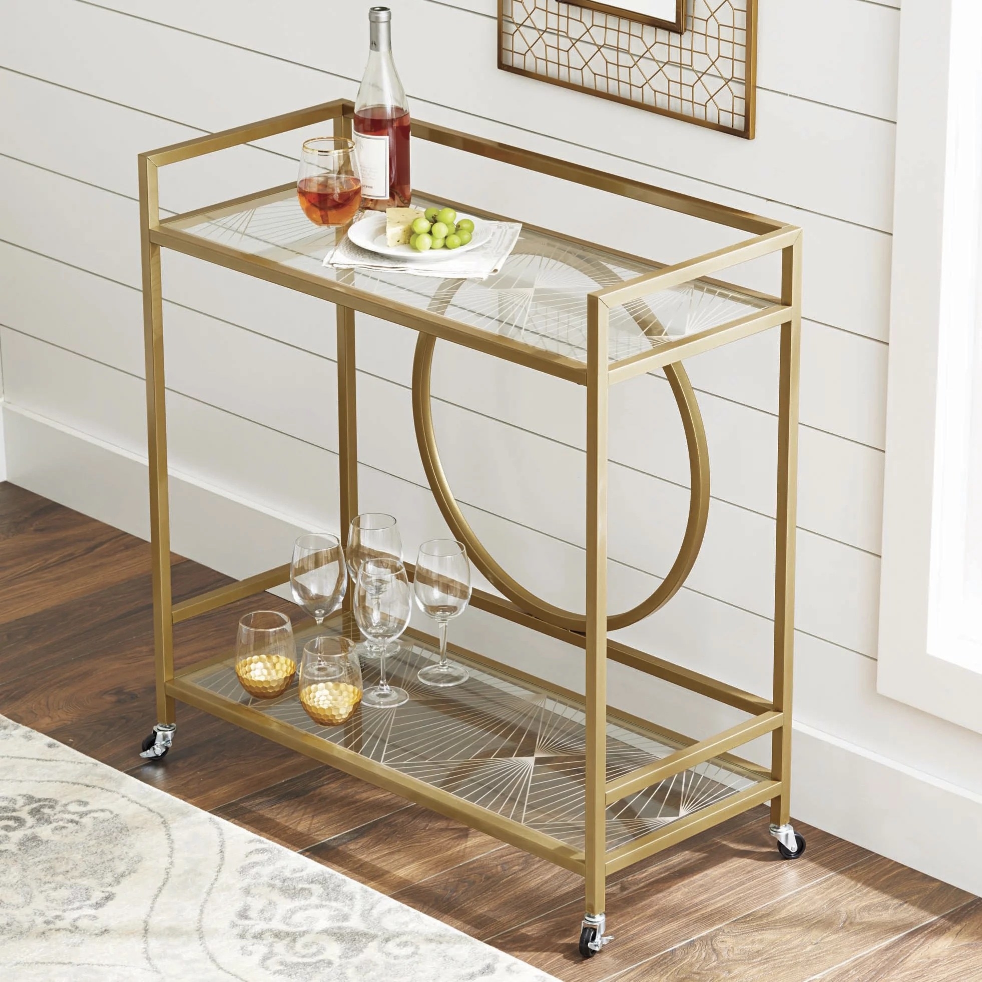 A gold bar cart with wine glasses on it anda bottle of wine and plate of cheese and grapes